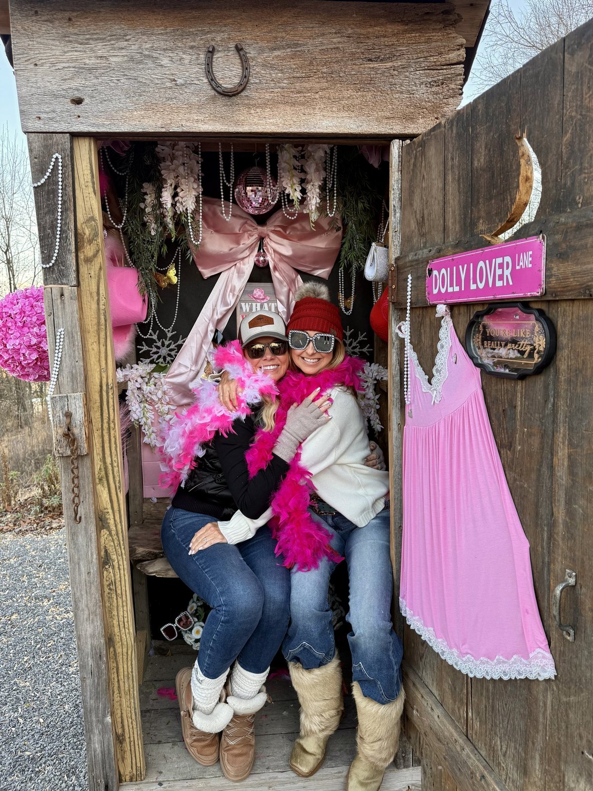 Two guests in cowgirl hats and pink feather boas posing inside Dolly’s Closet, beside a pink dress and a Dolly Lover sign, on the Sanctuary Treehouse Resort scavenger hunt